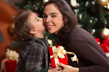 Fototapeta na wymiar Portrait of mother and son celebrating Christmas or New Year. Cute little boy is kissing his mother and giving her a Christmas present. Cheerful young mother is smiling and looking at camera.
