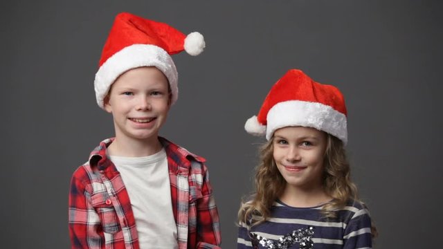 Little girl and boy in christmas caps looking at each other and then to the camera and smiling, gray background