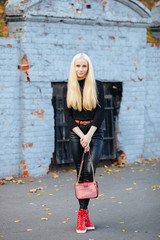 Young stylish sporty blond beautiful teen girl in black posing at park on a warm fall day against old cobalt painted brick wall background. Red clutch, vintage sneakers, belt, black outfit.