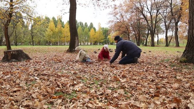 Father playing with daughter and son in autumn park on the fallen leaves, they are sit on the leaves and throw leaves at each other
