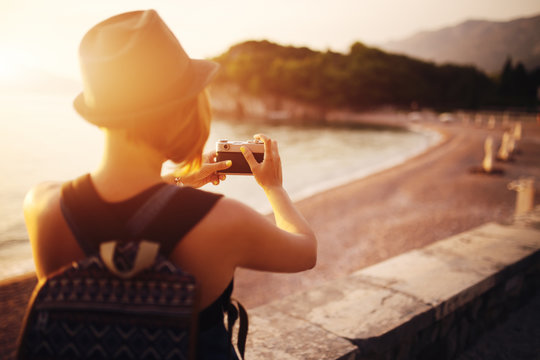 Woman traveler with camera taking photo near sea at sunset
