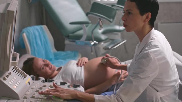 Pregnant woman lying on examination table and smiling at female doctor as she performing ultrasound scanning 