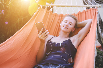 Summer day, a girl lying in a hammock with eyes closed and enjoying the music.Girl listening to music with headphones while lying in the orange hammock.Film effect.Girl using gadget.