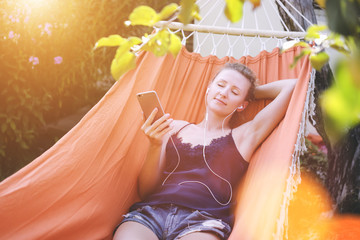 Front view of the girl lying in a hammock, holding a smart phone and listening to music.Film effect, blurred background.Girl using gadget.Girl using gadget.