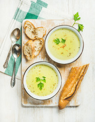 Green vegetable soup with parsley and baguette on white shabby wooden board over white painted wooden background, top view, vertical composition