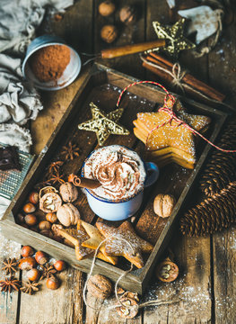 Mug of hot chocolate with whipped cream, cocoa powder, cinnamon, gingerbread cookies, nuts in wooden tray with Christmas decoration toys and pine cones over rustic wooden background, selective focus