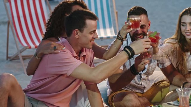 Four young men and women sitting together on the beach, toasting and drinking fruity cocktails while having picnic together in the evening