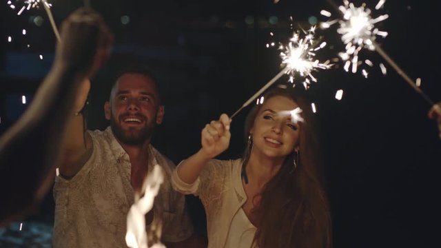 Young people celebrating something on the beach at night with firework sparklers, one couple kissing and hugging