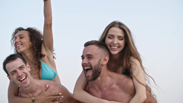 Two shirtless young men giving piggyback ride to their joyous girlfriend on the beach at summer day