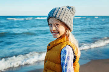 Portrait of smiling blonde white Caucasian child kid girl  with long hair, wearing yellow jacket gilet and grey hat, on beach at sunset looking in camera, autumn fall, happy lifestyle childhood