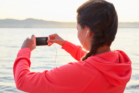 Active young woman listening to music during workout. Runner rests from routine exercise and makes photos. Happy jogger relaxing at the beach after run and looking at the sea. Back view