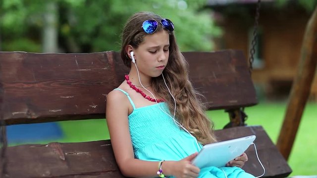 Pretty girl sits on swing bench and communicates via tablet computer. Beautiful girl with tablet computer sits on swing bench in garden. Attractive girl with tablet pc communicates through skype