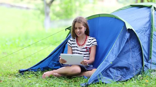 Pretty girl sits on grass near tourist tent and communicates via tablet computer. Girl with tablet pc sits on green grass near tourist tent. Pretty girl with tablet pc communicates through skype