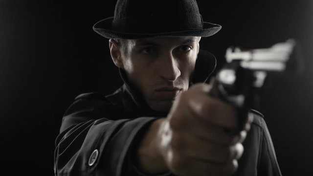 Portrait of a gangster in a hat and a black cloak threatens with weapon. Mafioso aiming a gun at the camera
