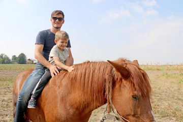 Strong handsome father in fashionable sunglasses with his small blond cute son walks with their horse in the field.