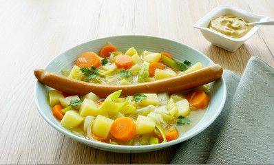 
Closeup of a vegetable stew with a large Frankfurter sausage on top and small bowl filled with mustard on light brown wooden background.  