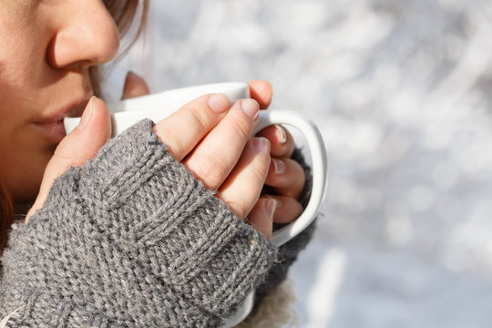 Woman in gray knitted mittens drinking from white cup at the bac