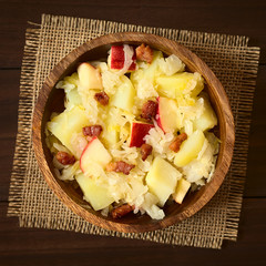 Potato, sauerkraut and apple salad with fried bacon served in wooden bowl, photographed overhead on dark wood with natural light