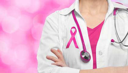 Female doctor with pink ribbon on chest against color background. Oncology and breast cancer concept.