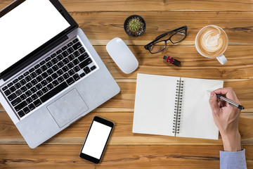 Young business man hands with pen writing leather notebook on desk table with blank screen laptop, blank screen smartphone, mouse, flahdrive, glasses and cup of coffee. Top view business concept.