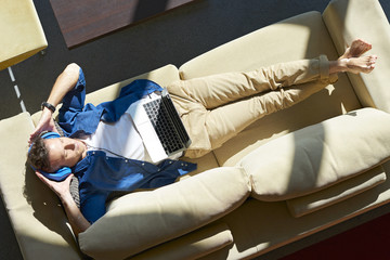 Directly above shot of a middle aged casual man lying on couch at home and  listening music with headphone on laptop while relaxing after work.