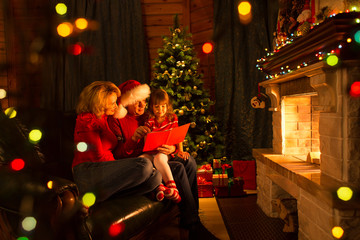 Happy family reading book at home by fireplace in warm and cozy living room on winter day...