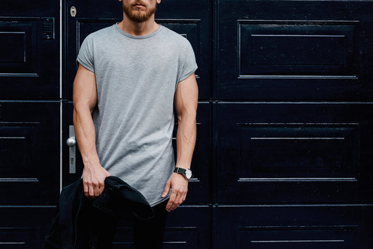 A cropped photo of a young muscular man wearing blank gray t-shirt while standing beside the dark entrance door on a street. Mock up.