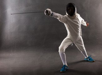 Boy wearing white fencing costume and black fencing mask standing with the sword practicing in...