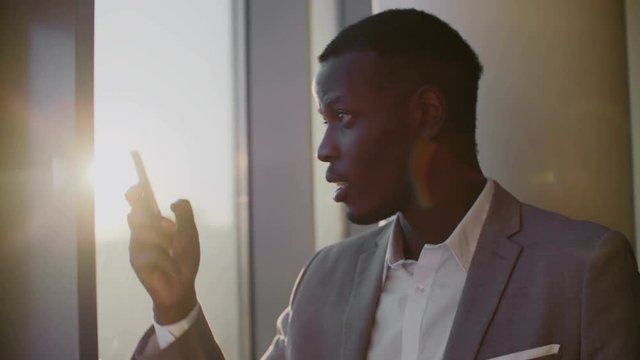 Young black man in suit using smartphone standing by window in ghetto office in the sun