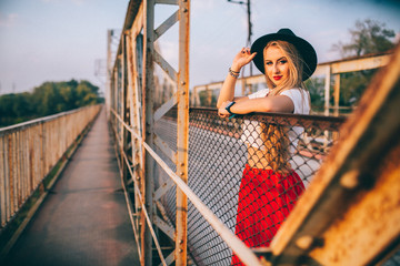 stylish portrait of a beautiful young woman on the bridge in a red skirt and black hat on a sunny day at sunset posing and smiling Life Style