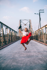 Outdoor lifestyle portrait of happy blonde young woman in stylish casual red dress (skirt) and black hat stay on bridge on the street. Girl throws hat and jump on the brige. Having fun . Long hair