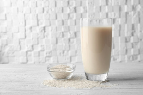 Glass of rice milk and bowl with grains on wooden table against white blurred background