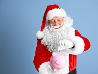 Santa Claus putting coin into piggy bank on blue background