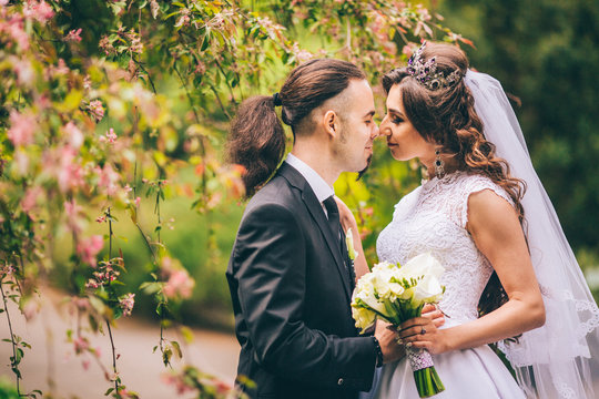 Happy elegant stylish groom with his gorgeous bride in the park. Man with long hair and piercing. Woman in beautiful gemstone crystal crown.