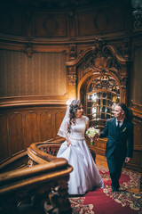 elegant gorgeous bride and stylish groom standing on wooden stairs in amazing old rich room. unusual wedding couple in retro style. luxury wedding concept. Man with long hair and piercing