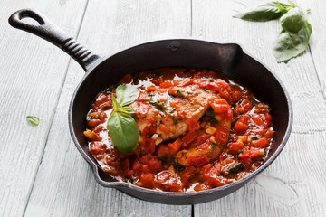 Stir-fried chicken breast in a sauce of tomatoes, garlic, basil and olive oil. Black cast-iron pan, light wooden table