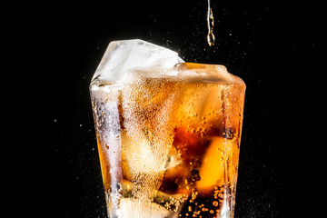Stream of soda being pouring into a glass with soda and ice cube on black background, closeup texture, splashing, splash, fizz 