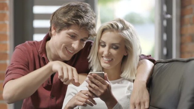 Attractive young couple watching smart phone together sitting on a couch in a living room at modern loft interior. Beautiful young hipster couple using phone at home.