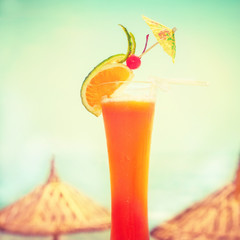Tequila sunrise cocktail with fruits  decoration at tropical ocean beach with umbrella. Vintage style, hipster colors image with copy space for party invitation text