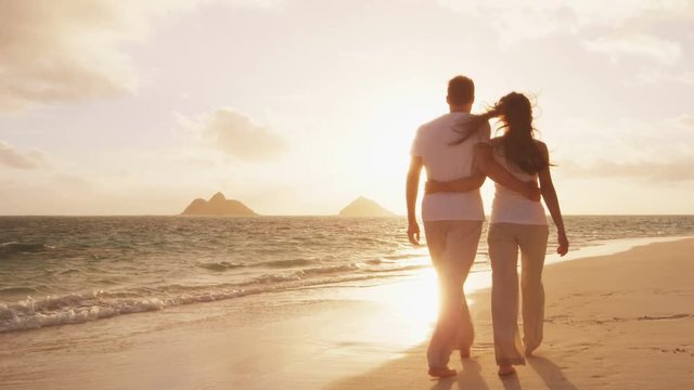 Beach sunset couple walking romantic on honeymoon in love. Sunrise romance young couple in elegant casual clothing walking together enjoying travel vacation holidays.