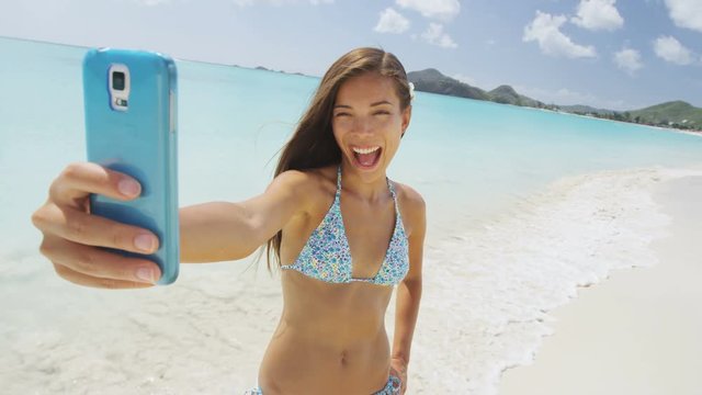 Selfie smart phone girl taking mobile phone photo on beach vacation during summer travel holidays. Sexy young bikini woman posing for camera having fun laughing on amazing Caribbean beach on Antigua.