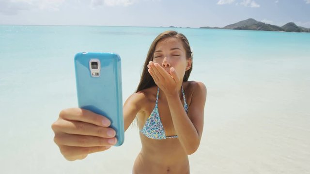Phone girl using smart phone on beach blowing kiss having fun on beach vacation during summer travel holidays taking a selfie photo or having video chat. Sexy young bikini woman posing for camera.