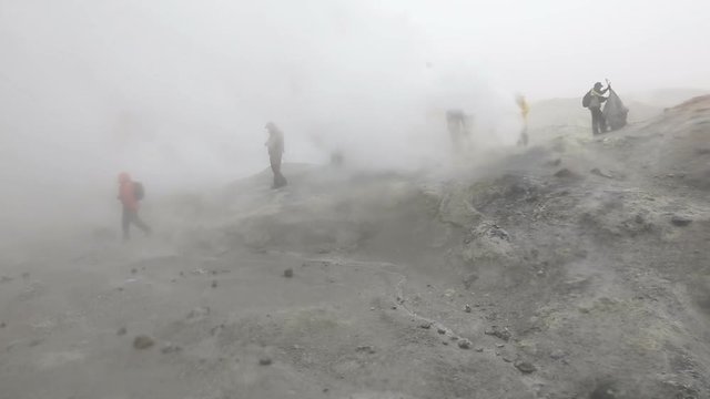 Hiking on the Kamchatka Peninsula - group of tourists walk on the smoking (steaming) fumarole field in crater active Mutnovsky Volcano in overcast rainy weather. Russian Far East, Kamchatka Region.