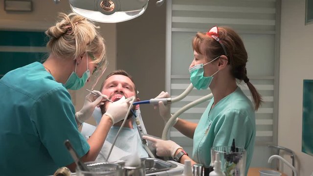 Dentist and assistant removing dental plaque from the teeth. Visit is in proffessional dental clinic. He is sitting on dental chair. He is young and has beard. Steadicam shot.
