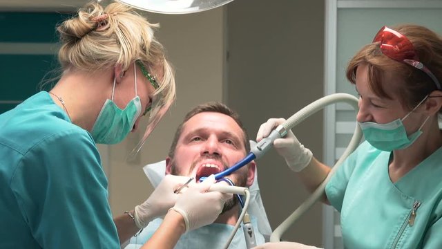 Dentist and assistant removing dental calculus from the teeth. Visit is in proffessional dental clinic. He is sitting on dental chair. He is young and has beard. Steadicam shot.
