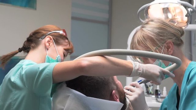 Female dentist and assistant removing dental calculus from the teeth. Visit is in proffessional dental clinic. He is sitting on dental chair. He is young and has beard. Steadicam shot.

