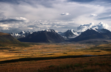 A highland river valley with yellow grass on a background of snow covered mountains and glaciers under clouds and blue sky, Plateau Ukok, Altai mountains, Siberia, Russia