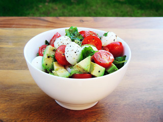 Fresh salad with avocado slices, cherry tomatoes and mozzarella. White bowl on a wooden table. High angle shot.