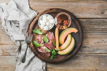 Fotobehang Assortiment Prosciutto di Parma, cantaloupe melon, mozzarella cheese in bowl, fresh basil leaves and glass of rose wine in round serving tray over rustic wooden background, top view, horizontal composition  © sonyakamoz
