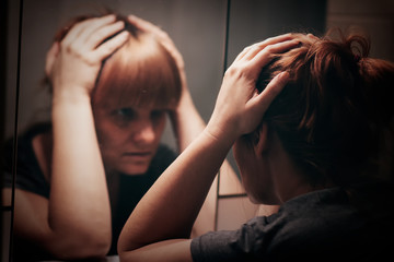 Sad and lonely woman holding her head and looking at her reflection in the mirror